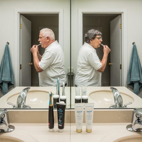 Ein älterer Mann putzt sich die Zähne, während ihm eine ältere Frau am Doppelwaschbecken im engen Raum begegnet.