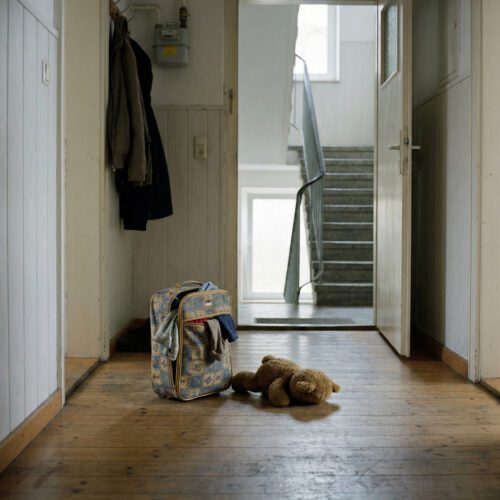 Ein halb gepackter Kinderkoffer steht verlassen neben einem Stofftier auf dem Boden vor einer offenen Wohnungstür.